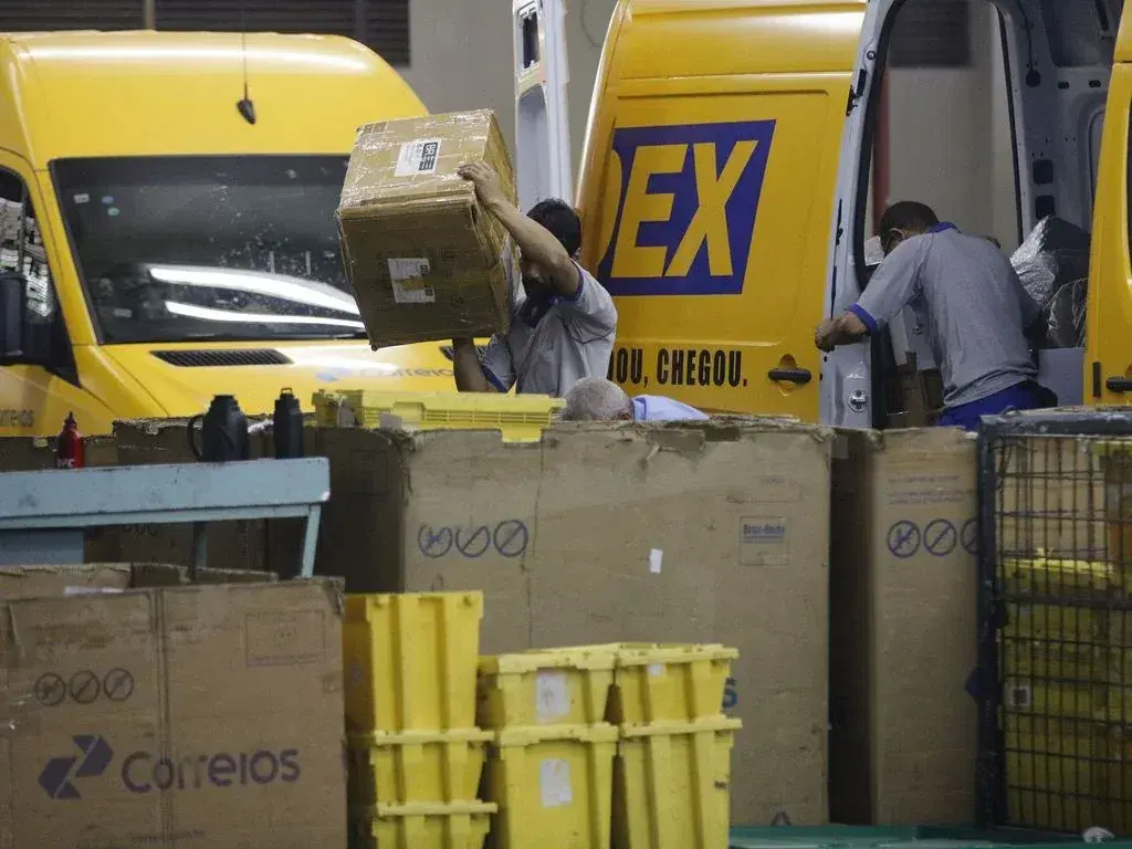 Funcionários movimentando encomendas junto a veículos dos Correios em centro de distribuição